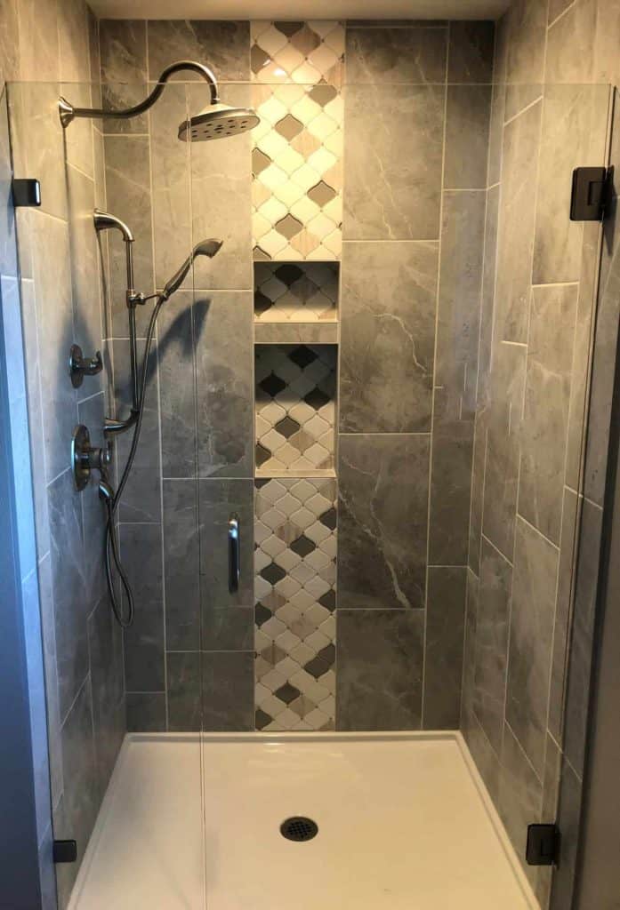 The new master bathroom shower includes a waterfall tile accent, a built-in niche with two shelves, clear glass pivot shower door, and a handheld shower arm along with a fixed shower head.